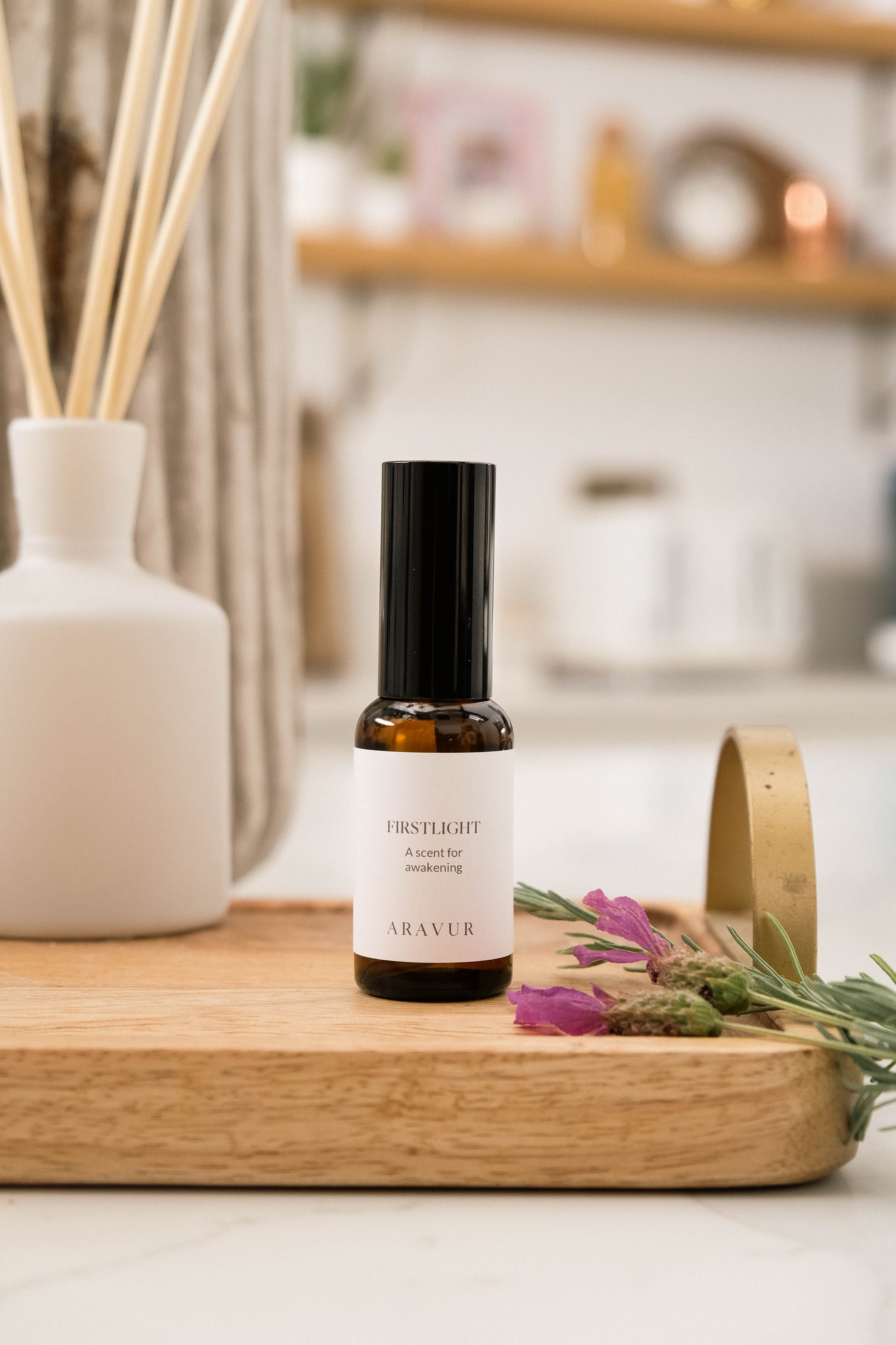 A bottle of 'Firstlight' room spray on a wooden tray with flowers and a diffuser in the background.