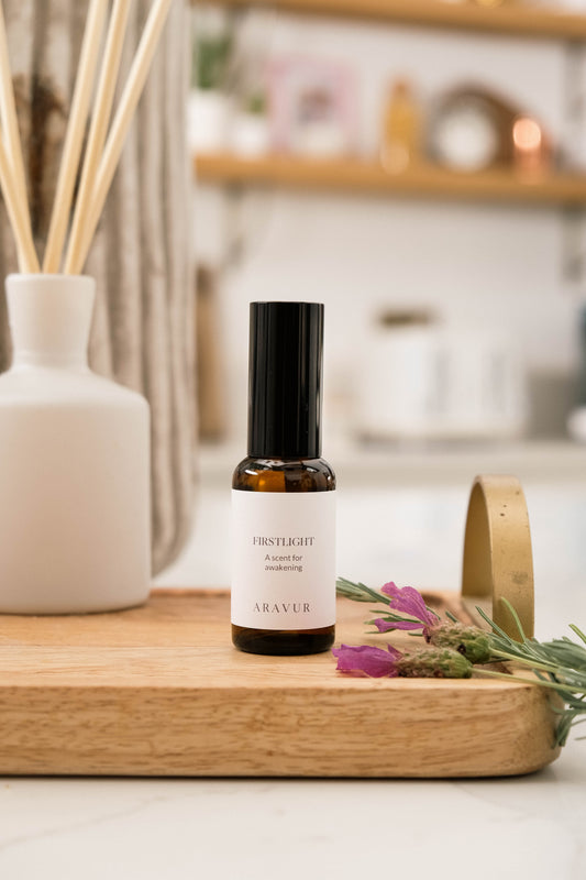 A bottle of 'Firstlight' room spray on a wooden tray with flowers and a diffuser in the background.